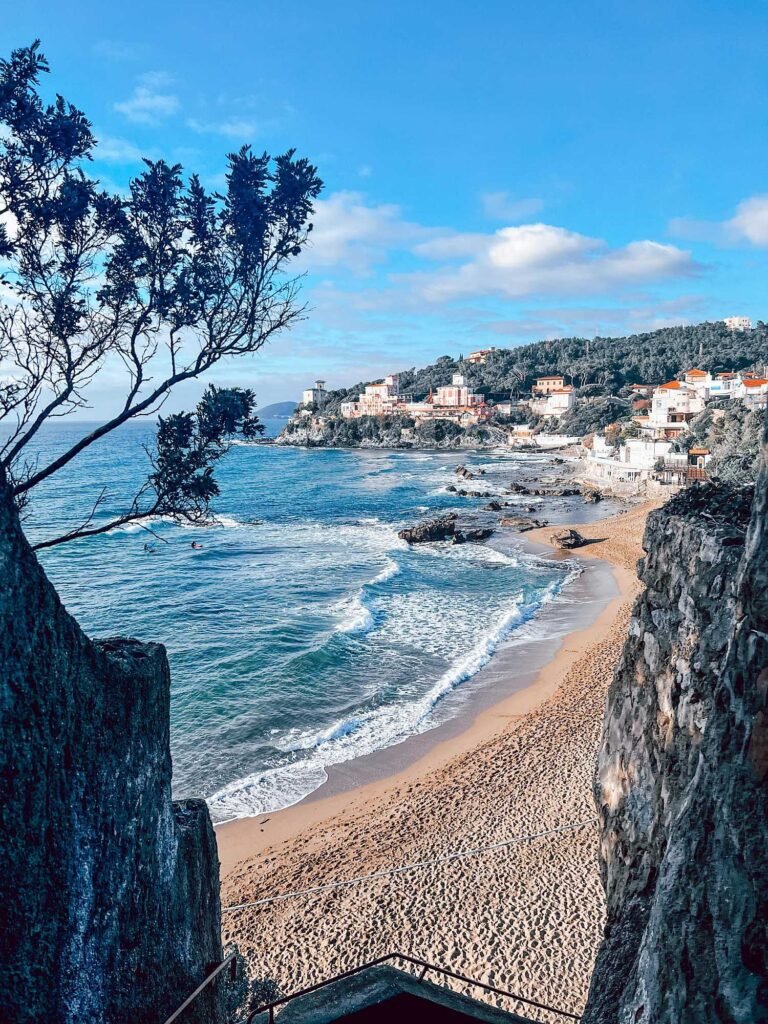 Spiaggia Baia del Quercetano a Castiglioncello con mare blu e case su scogliera, ideale per vacanze in Val di Cornia.